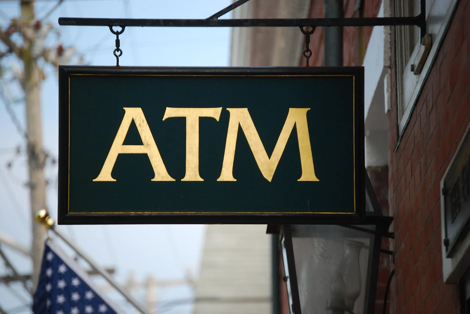 A vintage hanging ATM sign on a street corner beside a US flag